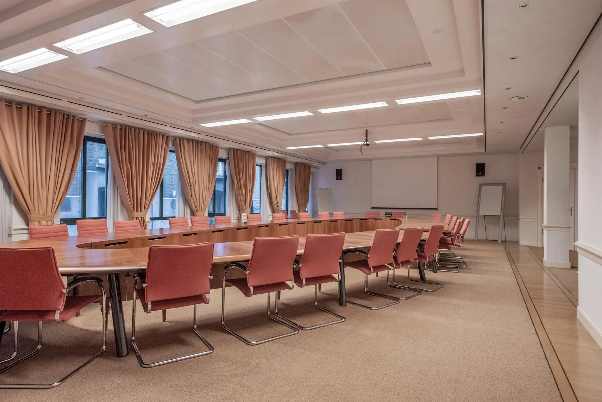 Ruime vergaderzaal aan de Doelenstraat met lange ovale tafel, roze stoelen en grote ramen met beige gordijnen.