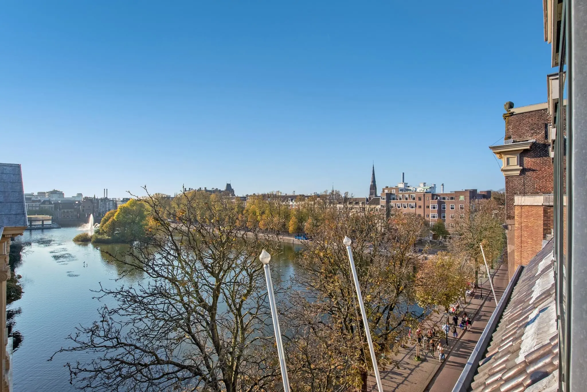 Uitzicht vanaf een hoog gebouw aan de Doelenstraat op een waterpartij, bomen in herfstkleuren en een stadssilhouet onder een heldere blauwe lucht.
