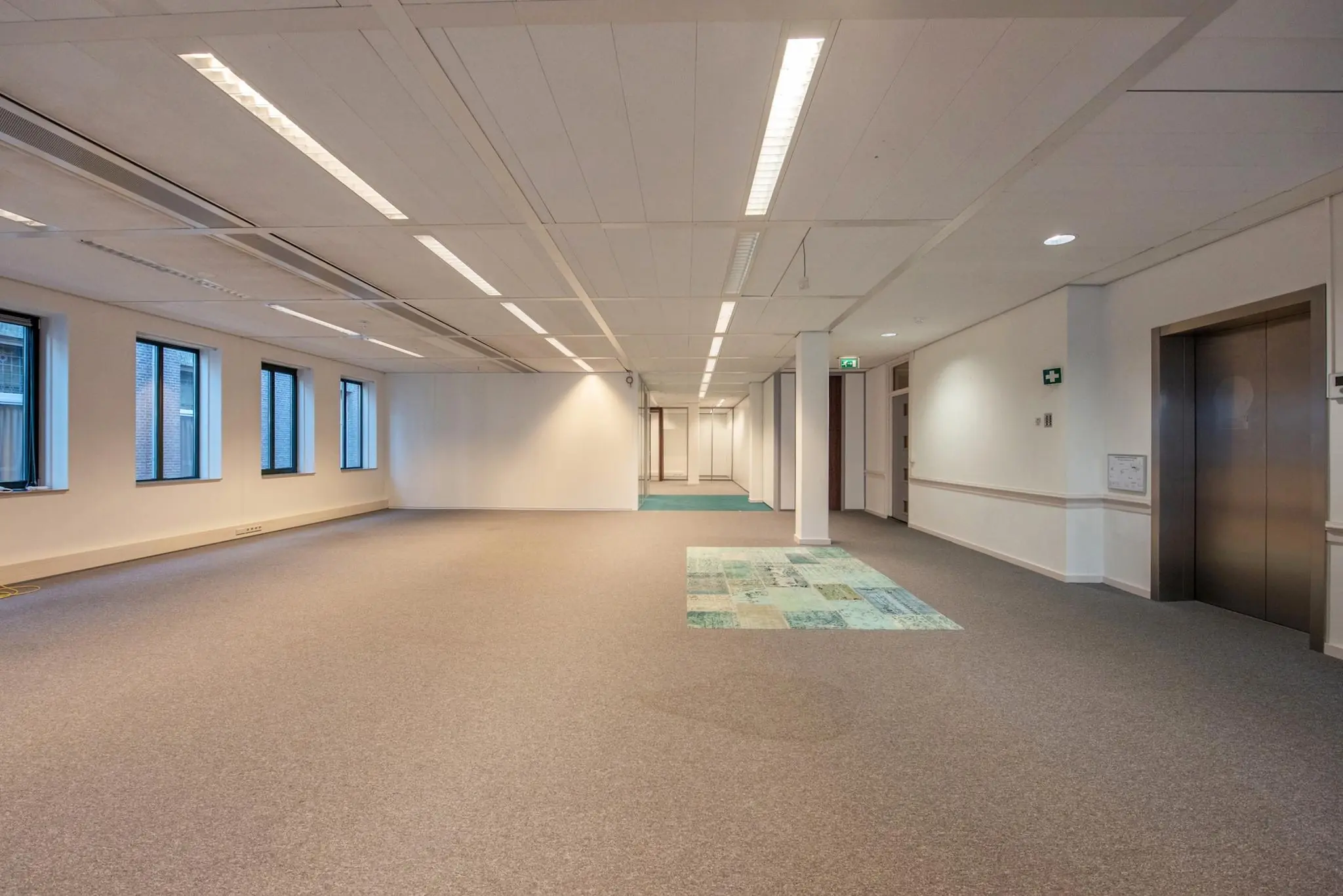 Leeg kantoorruimte met grijze vloerbedekking, ramen aan de linkerkant en een lift aan de rechterzijde aan de Doelenstraat.
