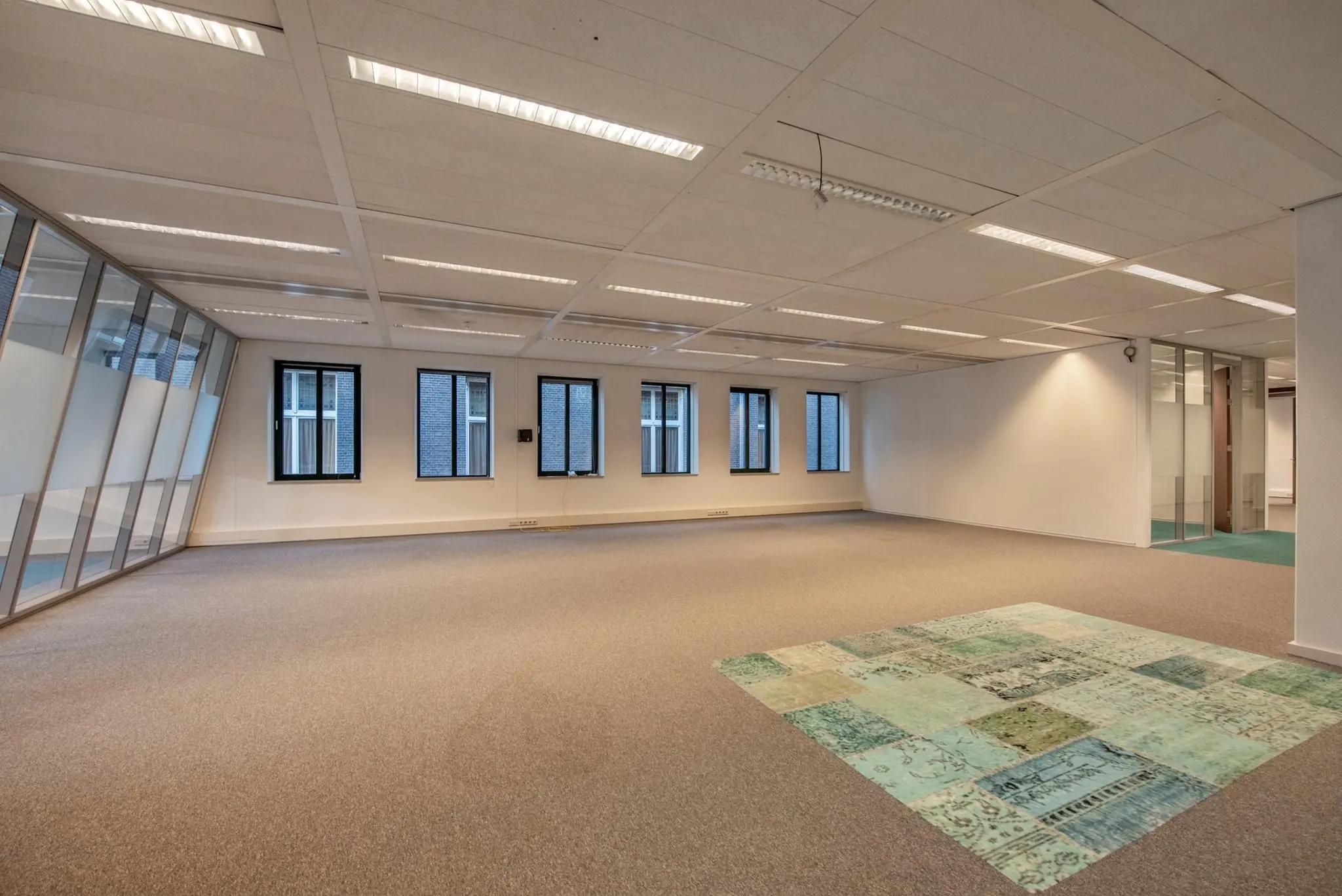 Leeg kantoorpand aan de Doelenstraat met grote ramen, systeemplafond en opvallend groen-blauw vloerkleed.