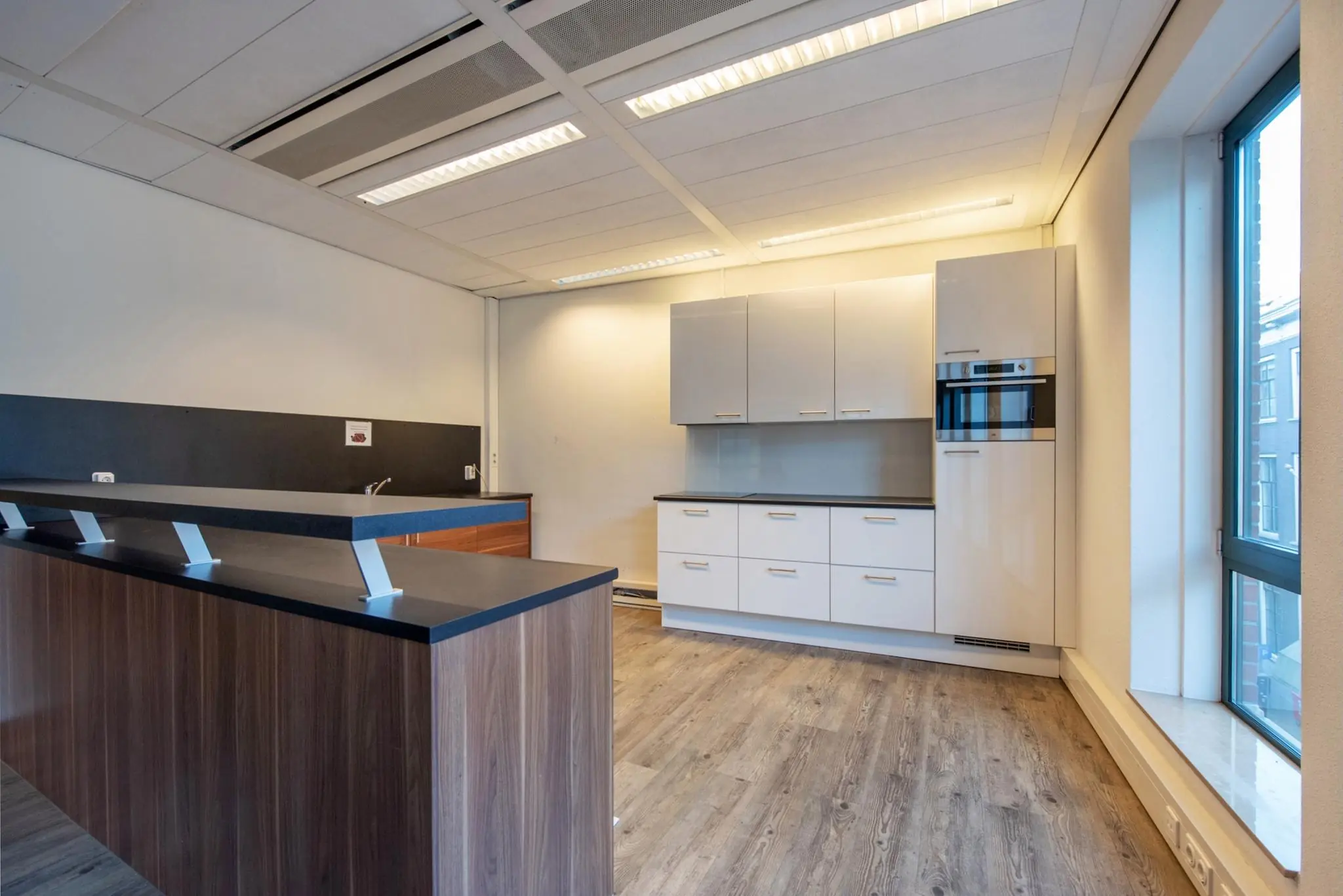 Moderne keuken in een kantoorruimte aan de Doelenstraat met houten vloer, witte kasten en groot raam.