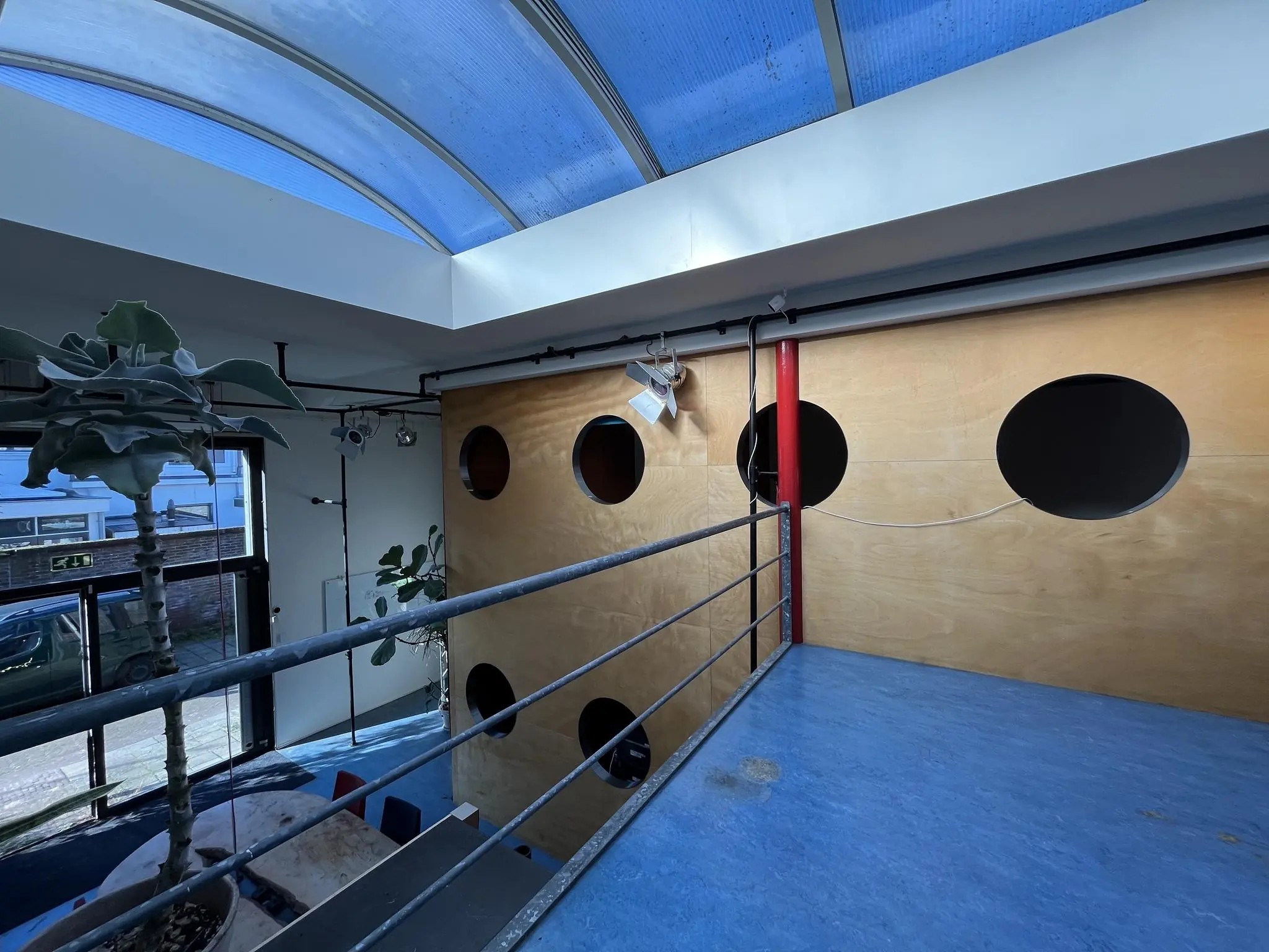 Binnenruimte aan de Kerkstraat met een blauwe vloer, houten wand met ronde openingen, glazen dak en zicht op benedenverdieping met planten en tafel.