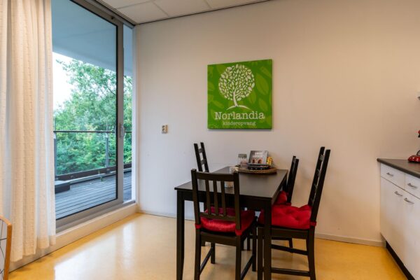 Kleine eethoek met vier stoelen en Norlandia kinderopvang-bord aan de muur, naast een glazen schuifdeur met uitzicht op groen.