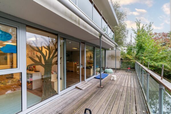 Houten balkon met glazen balustrade aan een kinderdagverblijf aan de Dedemsvaartweg, met zicht op speelruimte binnen en omringd door groen.