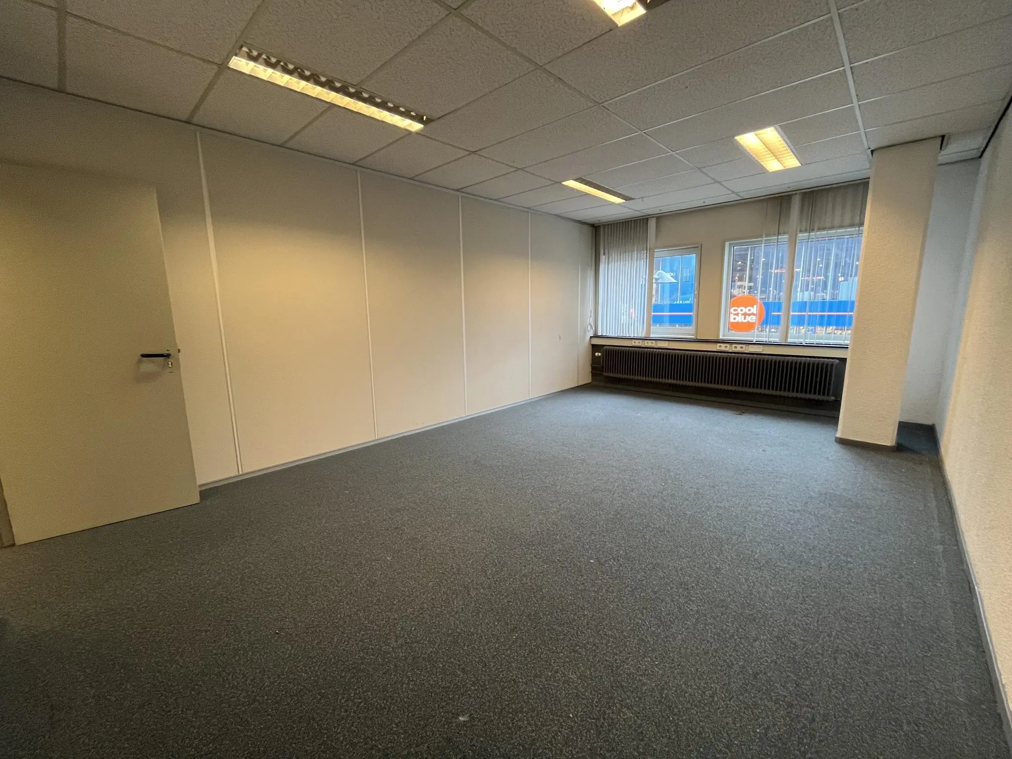 Leeg kantoorruimte aan de Hollantlaan met grijze vloerbedekking, systeemplafond en zicht op Coolblue-gebouw door de ramen.