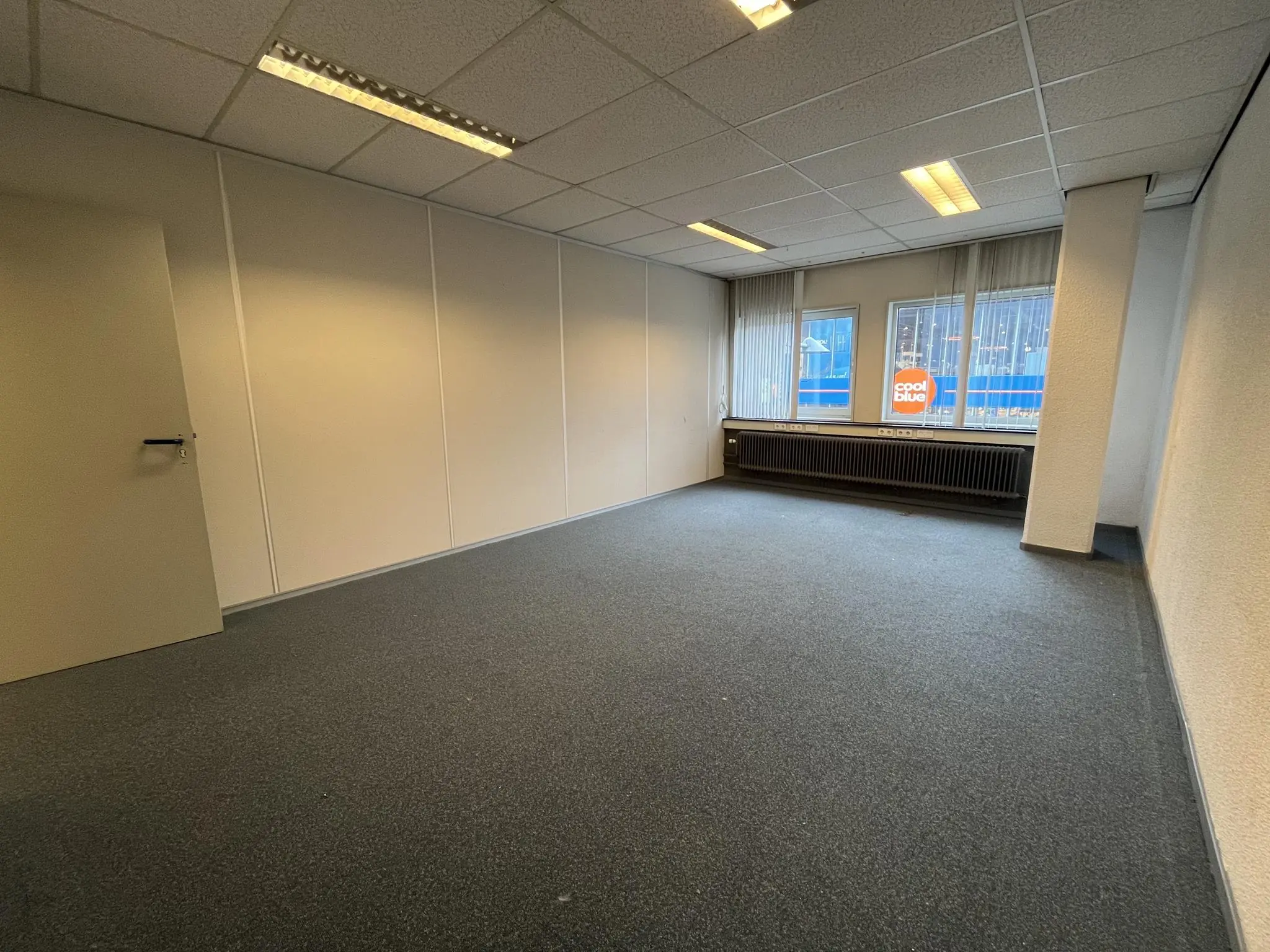 Lege kantoorruimte aan de Hollantlaan met tapijt, systeemplafond en zicht op een Coolblue-gebouw buiten.