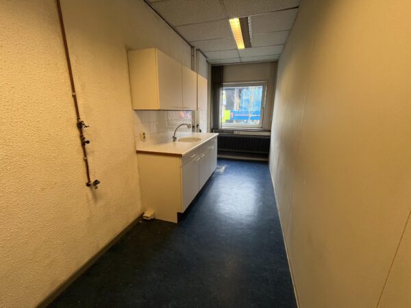 Smalle kitchenette aan de Hollantlaan met witte keukenkastjes, donkerblauwe vloer en een raam aan het einde.