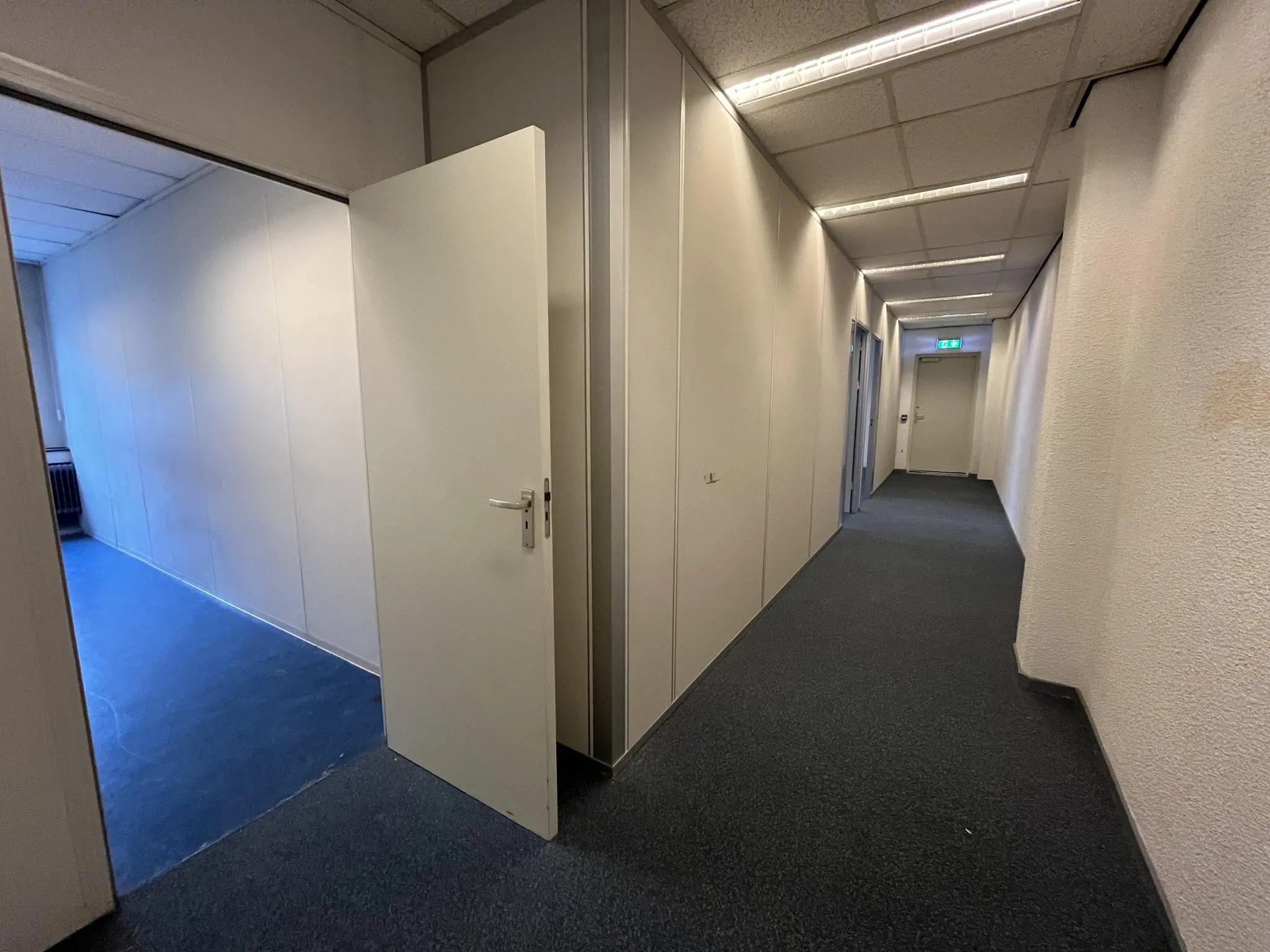 Gang in een kantoorpand aan de Hollantlaan met een openstaande deur naar een lege kamer met blauw vloerzeil.