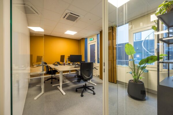 Modern kantoor met glazen wanden, vier werkplekken en een opvallende okergele accentmuur aan de Atoomweg.