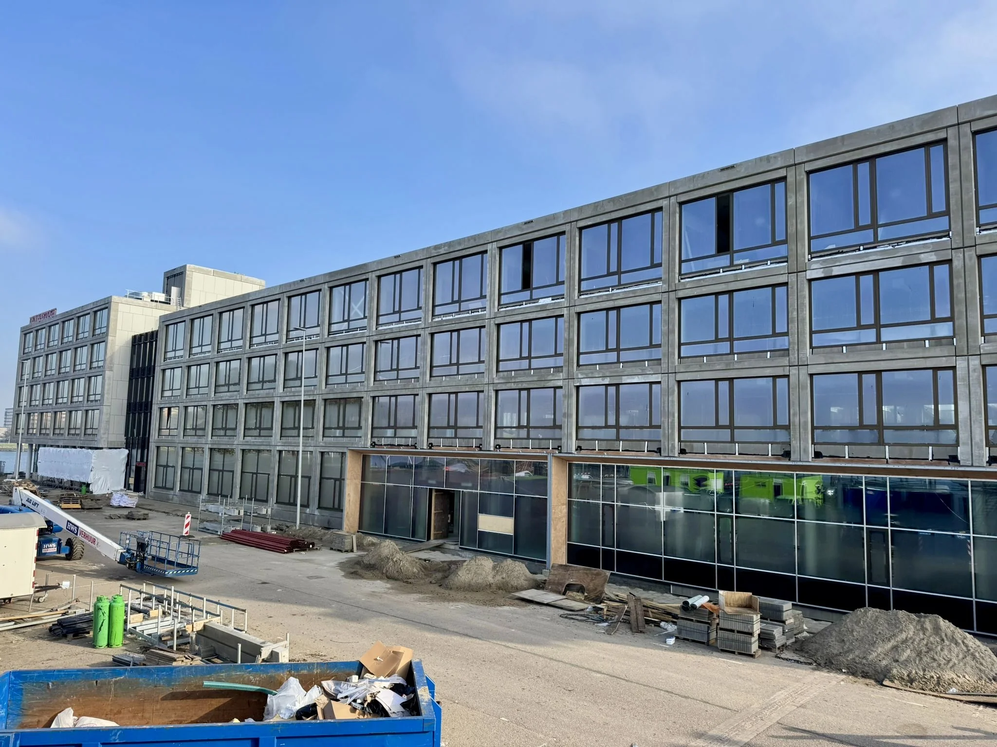 Groot modern gebouw aan de Sluisjesdijk in aanbouw, met glazen gevel en bouwmaterialen op de voorgrond.