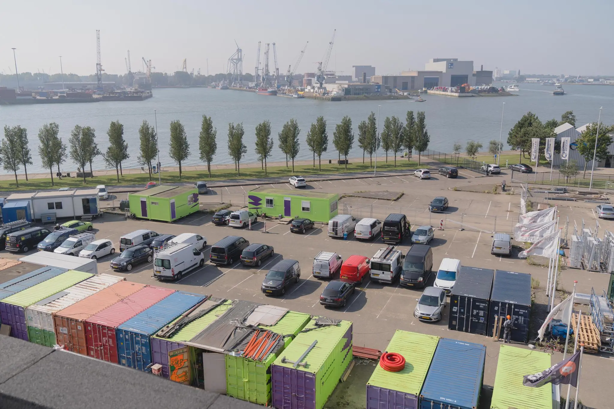 Parkeerterrein met felgekleurde containers en bouwunits aan de Sluisjesdijk, met uitzicht op de haven en kranen aan de overzijde van het water.