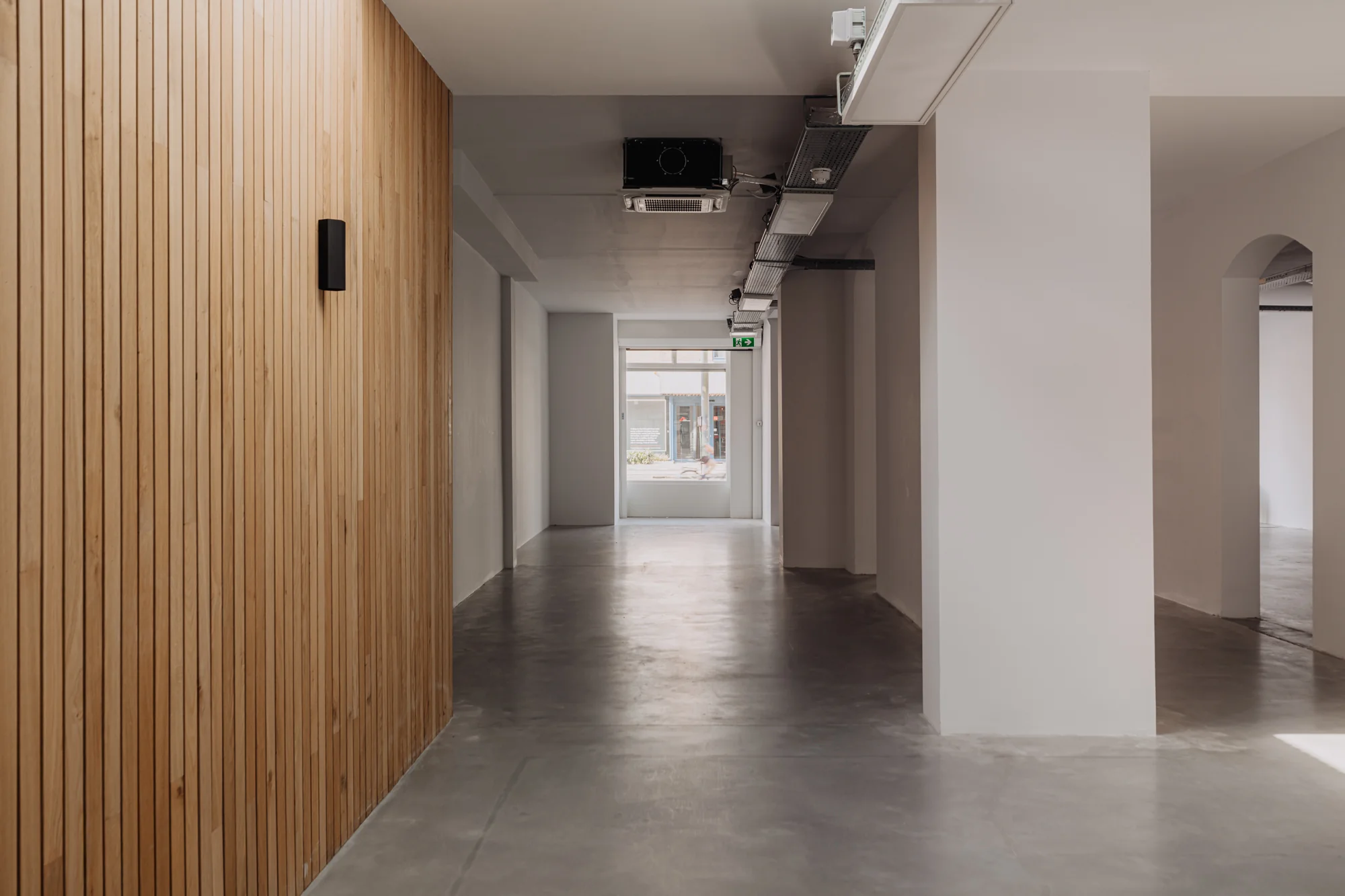 Moderne, lege gang met betonnen vloer, houten wandbekleding en zicht op een raam aan de Plantage Middenlaan.