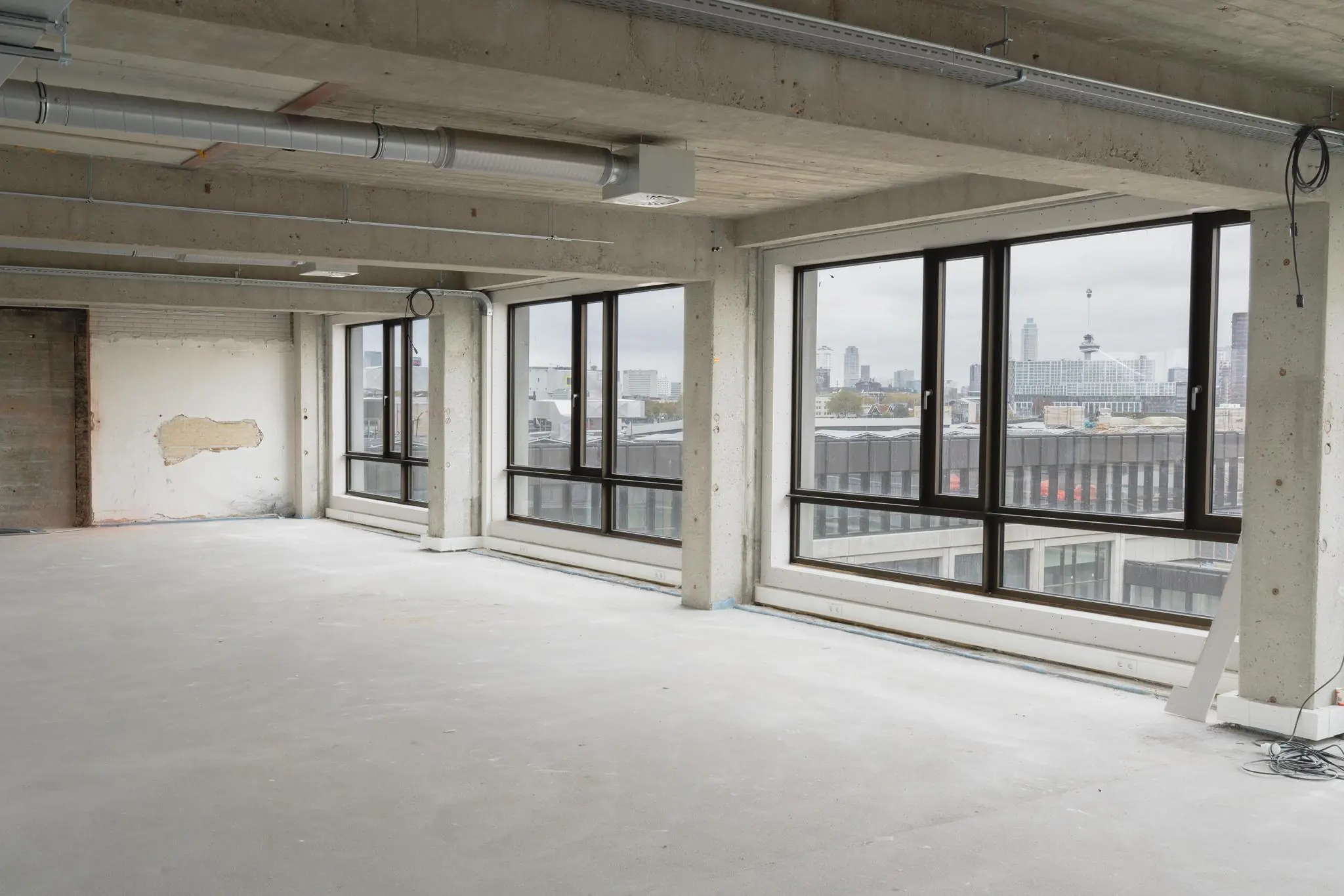 Leegstaand betonnen kantoorpand aan de Sluisjesdijk met grote ramen en uitzicht op de skyline van Rotterdam.