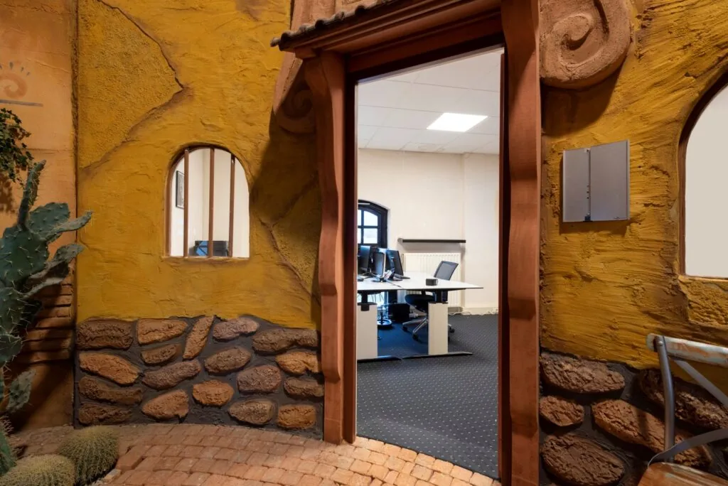 Kantoorruimte met bureaus en computers, gezien door een kleurrijke, zuidwesters gethematiseerde ingang met steen- en stucwerk.