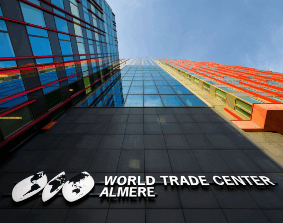 Voorgevel van het World Trade Center Almere met spiegelende ramen en rode accenten, gezien vanaf straatniveau.