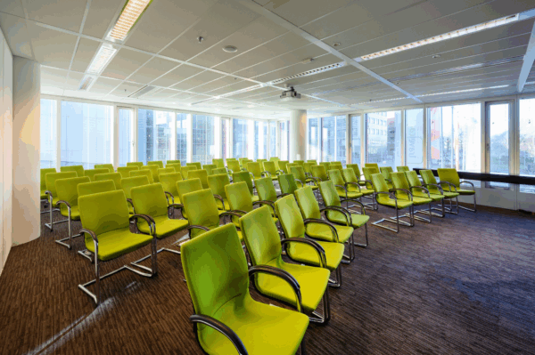 Vergaderruimte aan de P.J. Oudweg met rijen groene stoelen en grote ramen met uitzicht op moderne kantoorpanden.