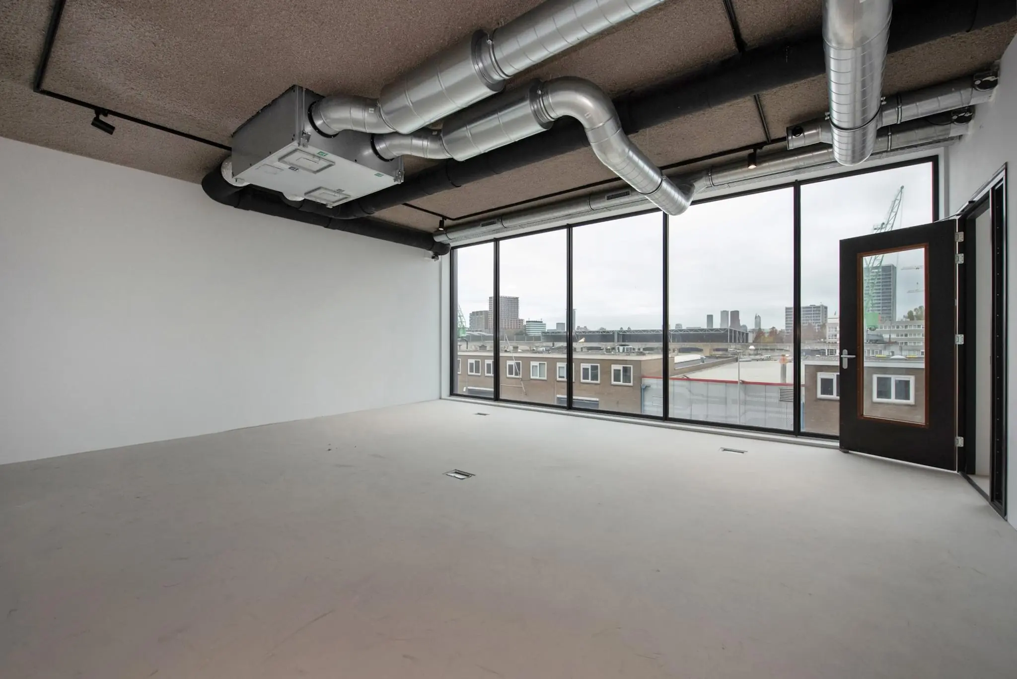 Licht industrieel kantoor met betonnen vloer, zichtbare ventilatiebuizen en grote ramen met uitzicht op de Saturnusstraat.