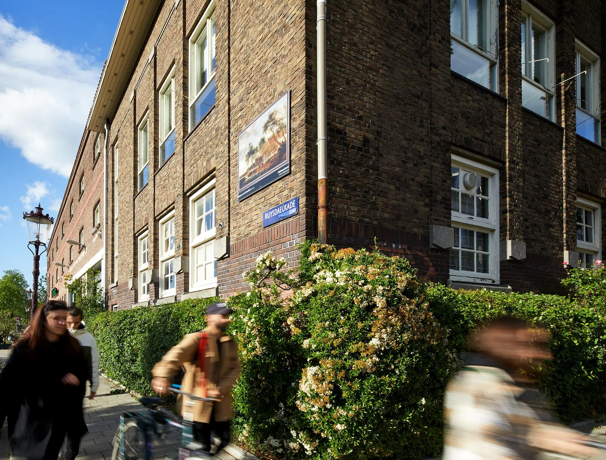 Straatbeeld van de Ruysdaelkade met een bakstenen gebouw, voorbijgangers en bloeiende struiken.