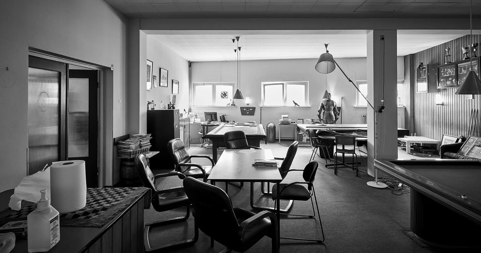 Zwart-witfoto van een ruime, nostalgisch ingerichte kantoorruimte aan de Ruysdaelkade met bureaus, zitjes en vitrines vol trofeeën.