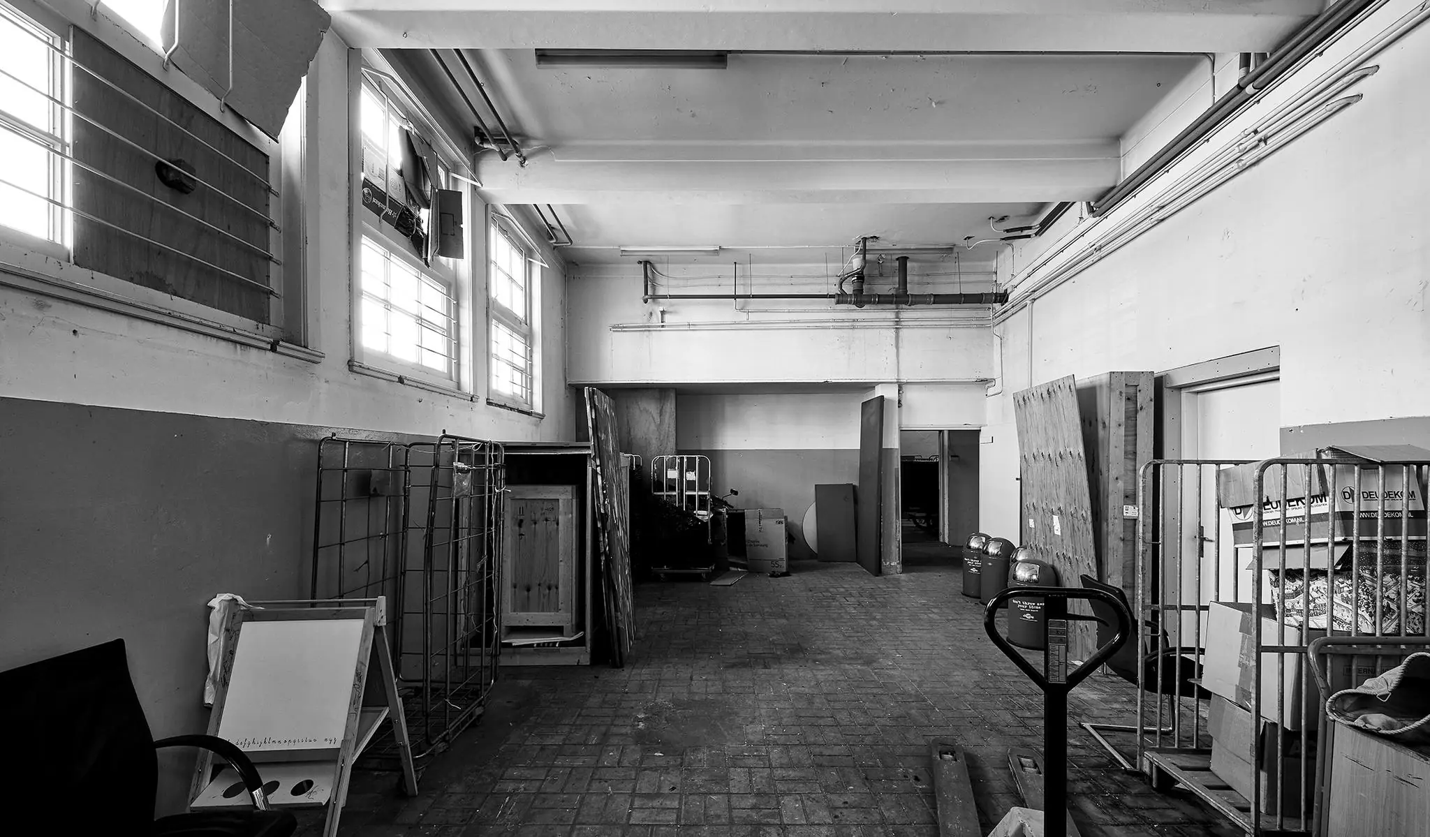 Zwart-witfoto van een lege, industriële opslagruimte aan de Ruysdaelkade met rekken, pallets en losse objecten.