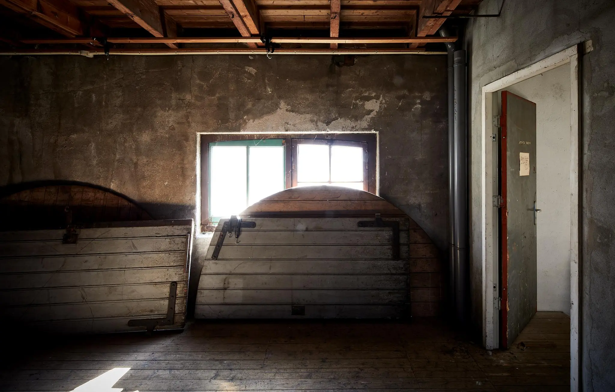 Een verlaten, stoffige ruimte aan de Ruysdaelkade met houten luiken, een klein raam en een deels open grijze deur.