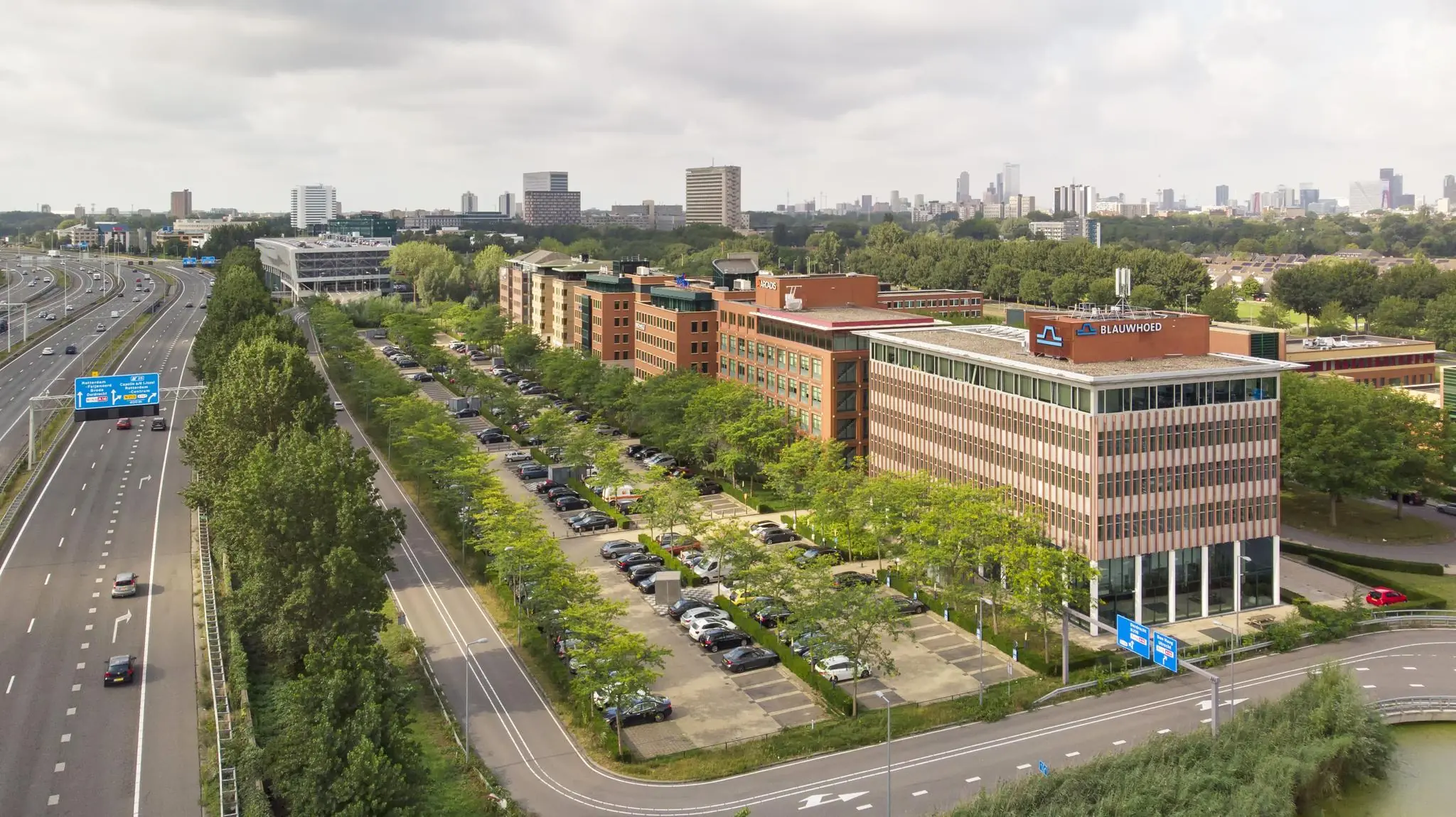 Luchtfoto van het kantoorgebouw Blaauwhoed aan de Lichtenauerlaan in Rotterdam, naast de A16 snelweg met parkeerplaats op de voorgrond.