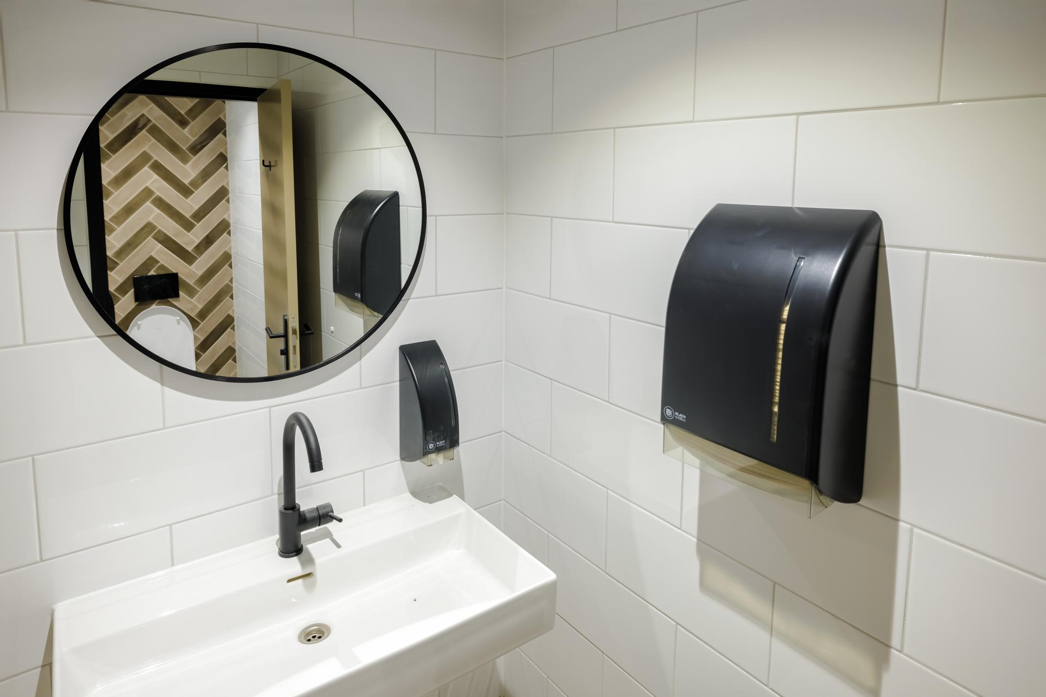 Moderne toiletruimte met witte wandtegels, een ronde spiegel, zwarte kraan en dispensers bij Hiridostraat 3.