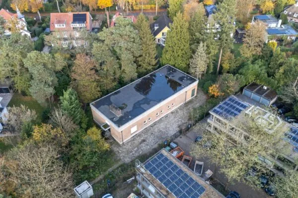 Luchtfoto van een rechthoekig gebouw aan de Rembrandtlaan, omringd door bomen en woningen met zonnepanelen.