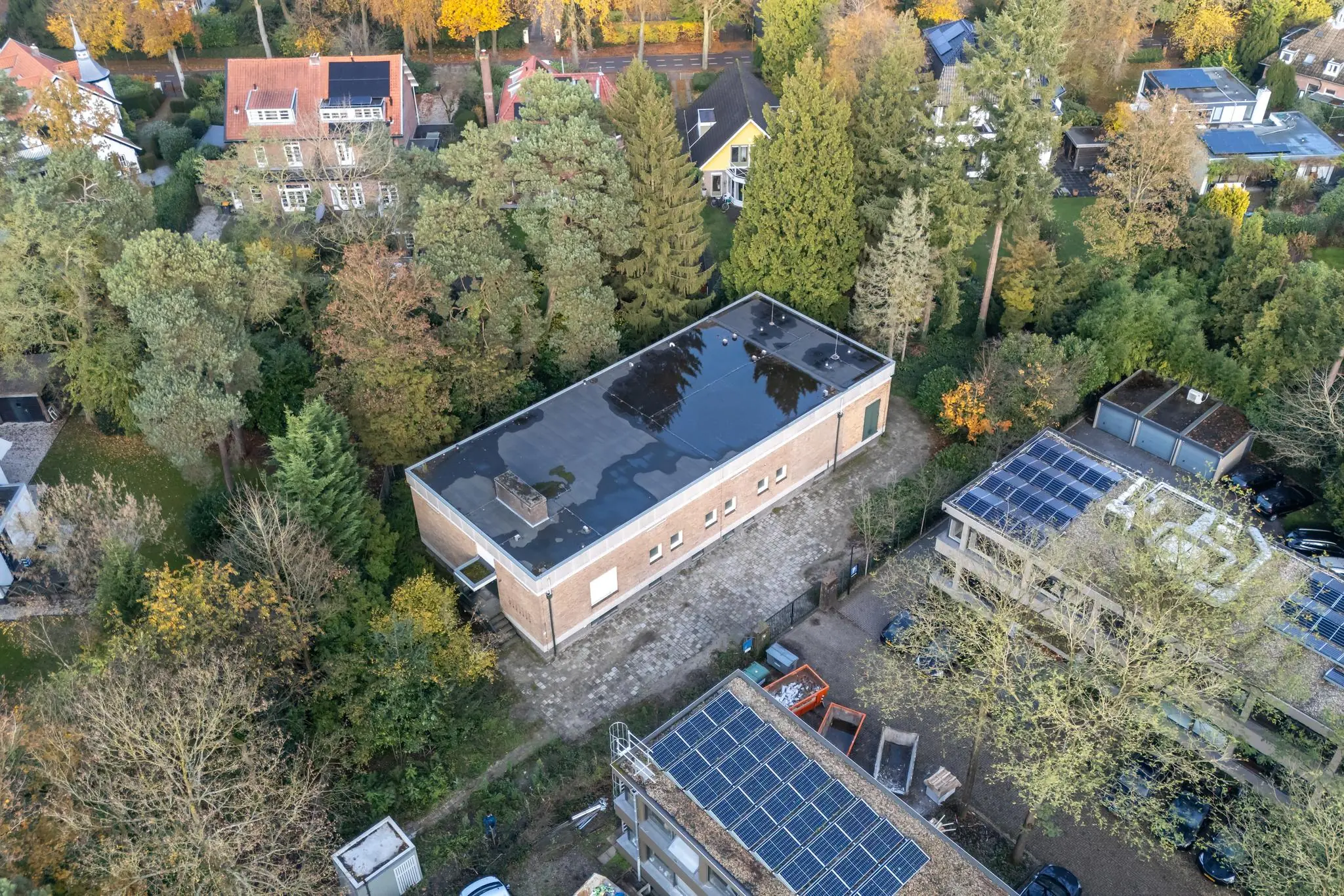Luchtfoto van een rechthoekig gebouw aan de Rembrandtlaan, omringd door bomen en woningen met zonnepanelen.