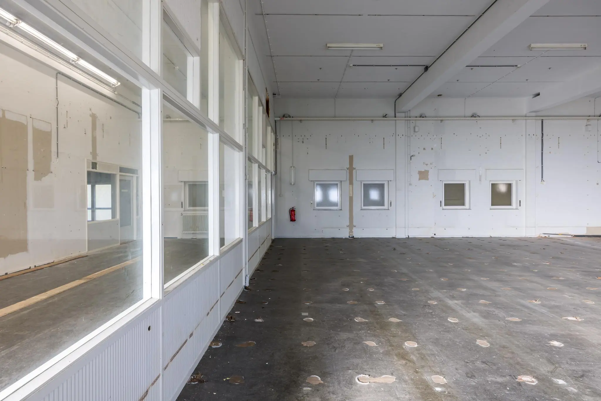 Leegstaande industriële ruimte aan de Rembrandtlaan met betonnen vloer, wandramen en zicht op aangrenzend kantoor.