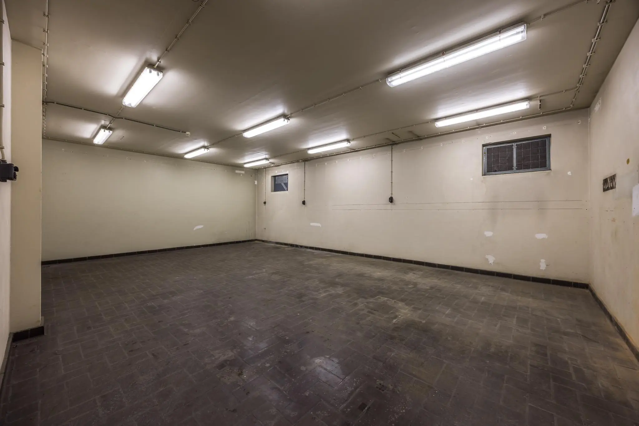 Lege, rechthoekige kelderruimte met tegelvloer, beige muren en tl-verlichting aan het plafond.