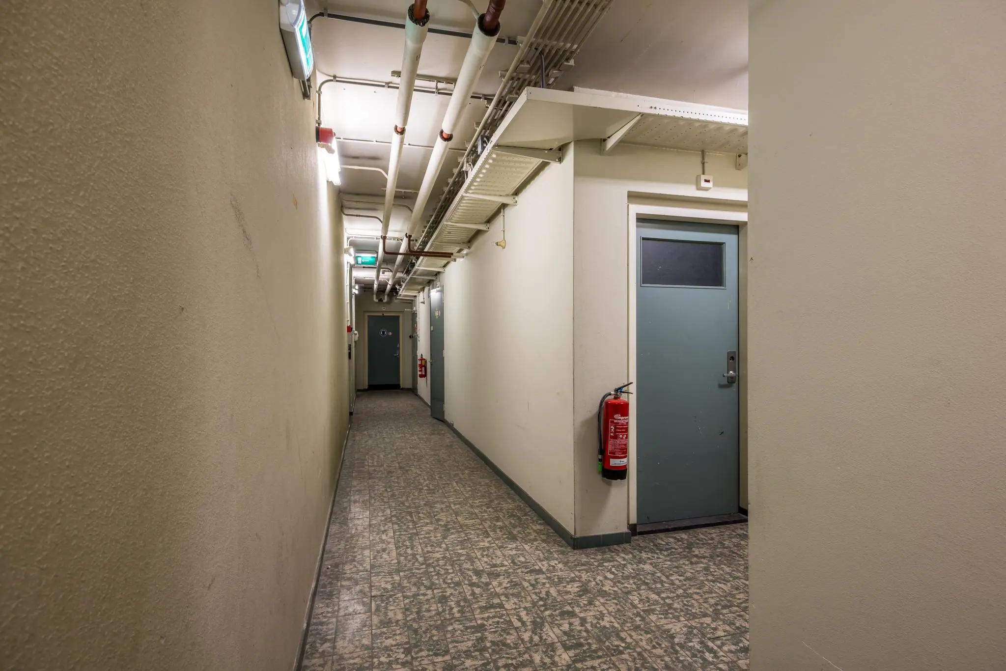 Langgerekte gang in een souterrain aan de Rembrandtlaan, met grijze deuren, leidingen aan het plafond en een brandblusser aan de muur.