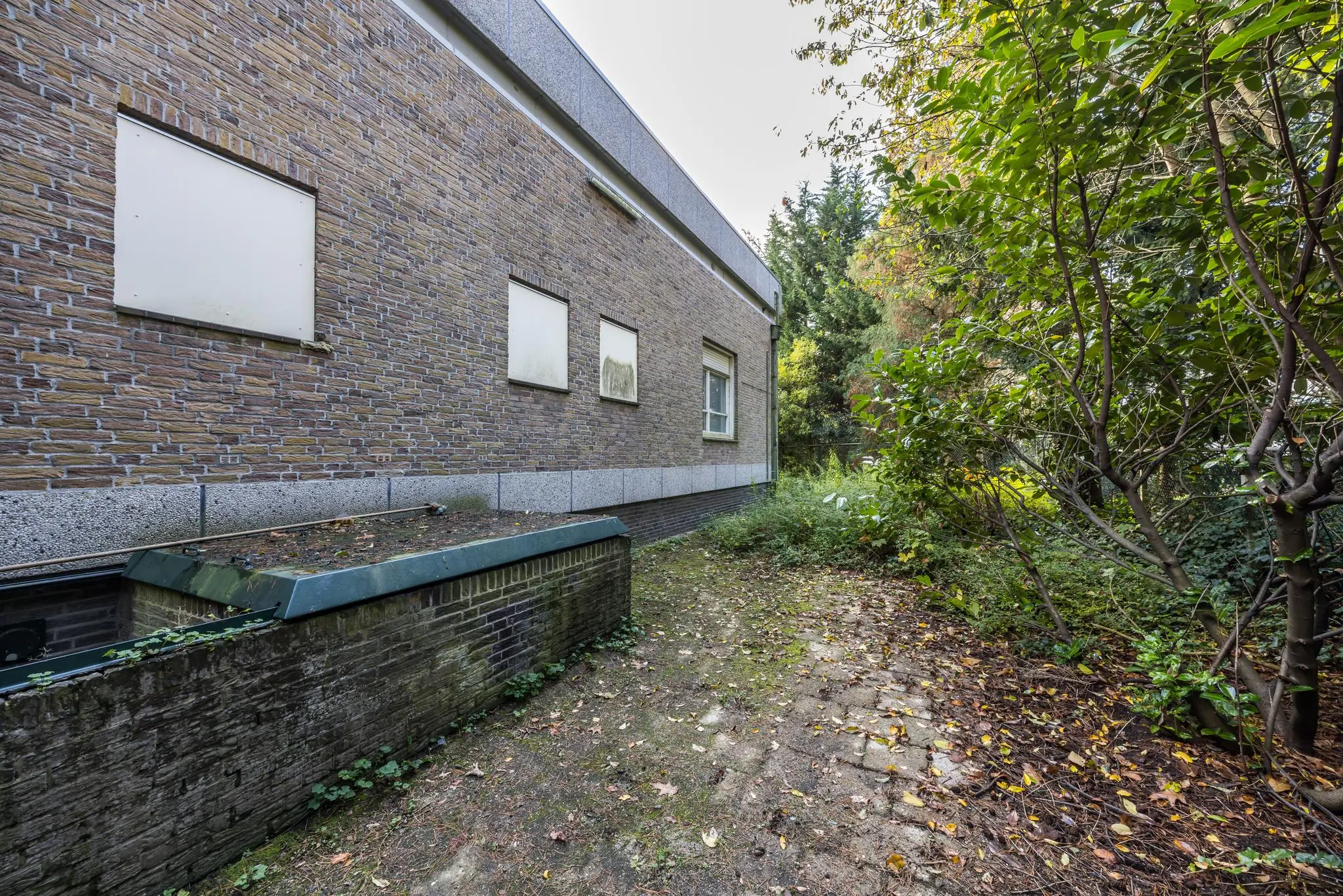 Zijaanzicht van een bakstenen gebouw aan de Rembrandtlaan met een smalle, deels overgroeide tuin en betegeld pad.