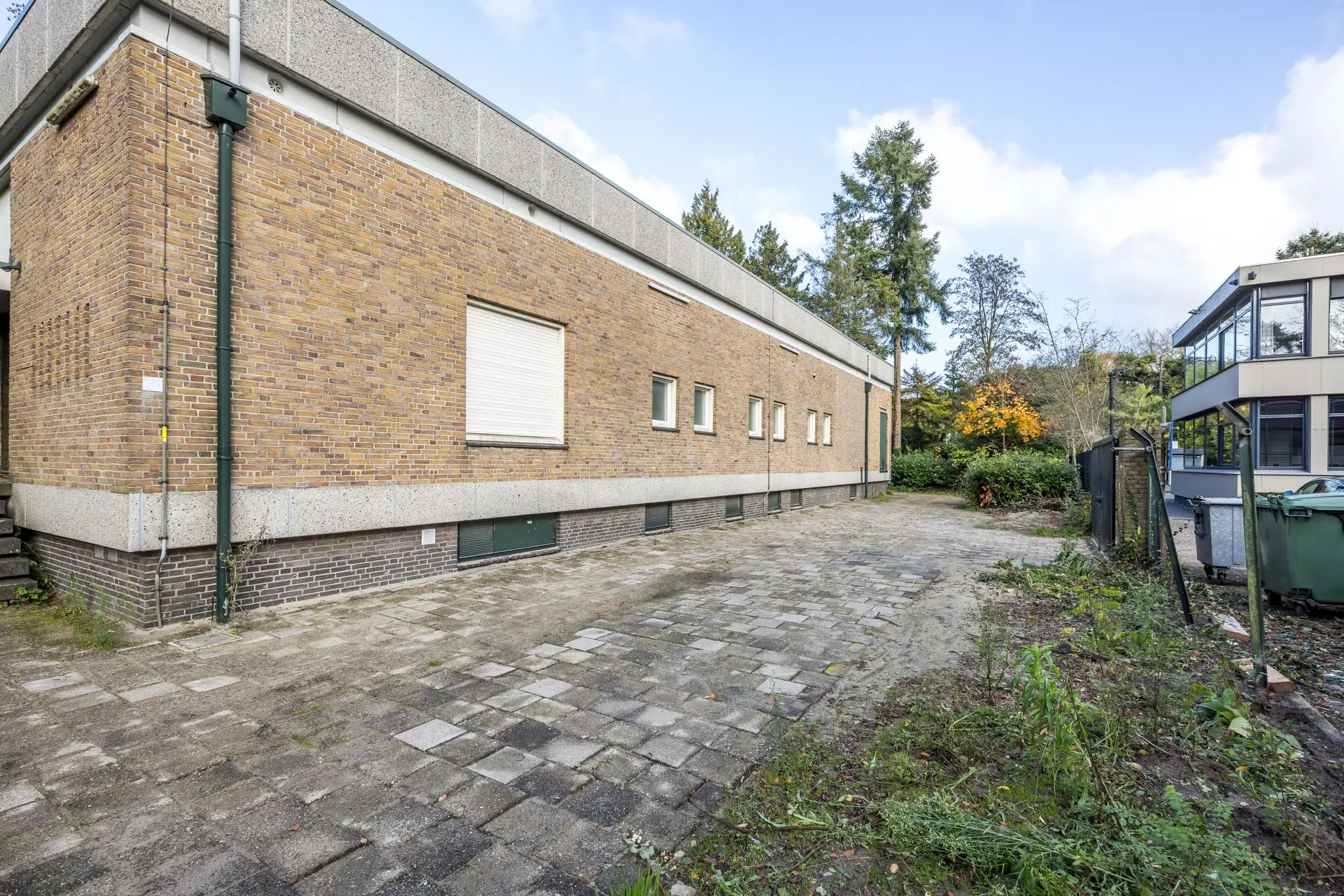Zijaanzicht van een laag, langwerpig bakstenen gebouw aan de Rembrandtlaan met bestrate oprit en omliggend groen.