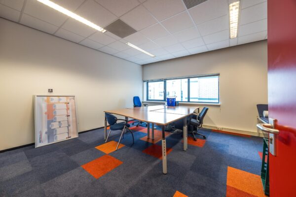 Kantoorruimte met bureaus, stoelen, kleurrijke vloerbedekking en een groot raam aan de achterzijde.