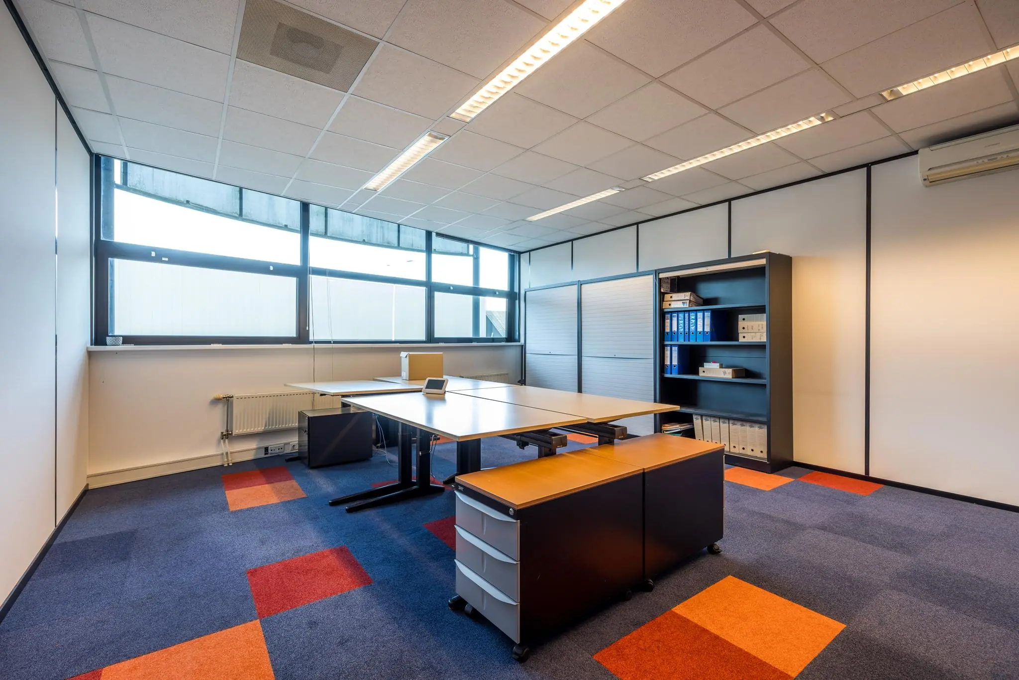 Kantoorruimte aan de Atoomweg met vergadertafel, archiefkasten en felgekleurd tapijt.