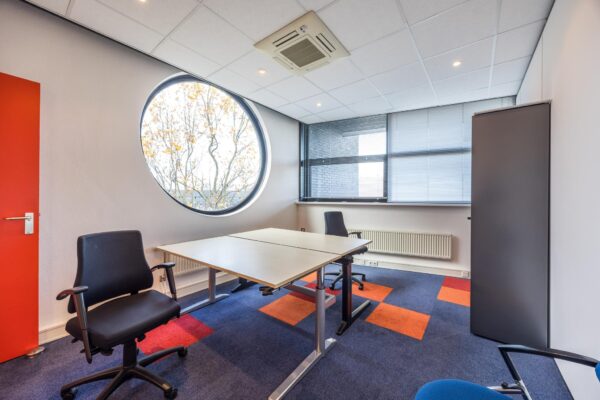 Modern kantoor met twee bureaus, kleurrijk tapijt en een groot rond raam met uitzicht op bomen.