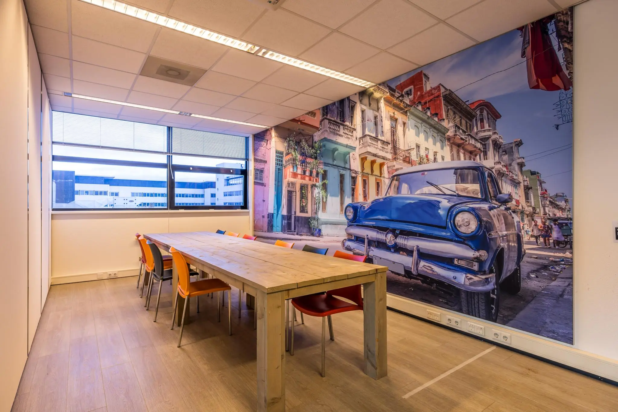 Vergaderruimte met een houten tafel, kleurrijke stoelen en een muurdecoratie van een klassieke straat in Havana met een blauwe oldtimer.