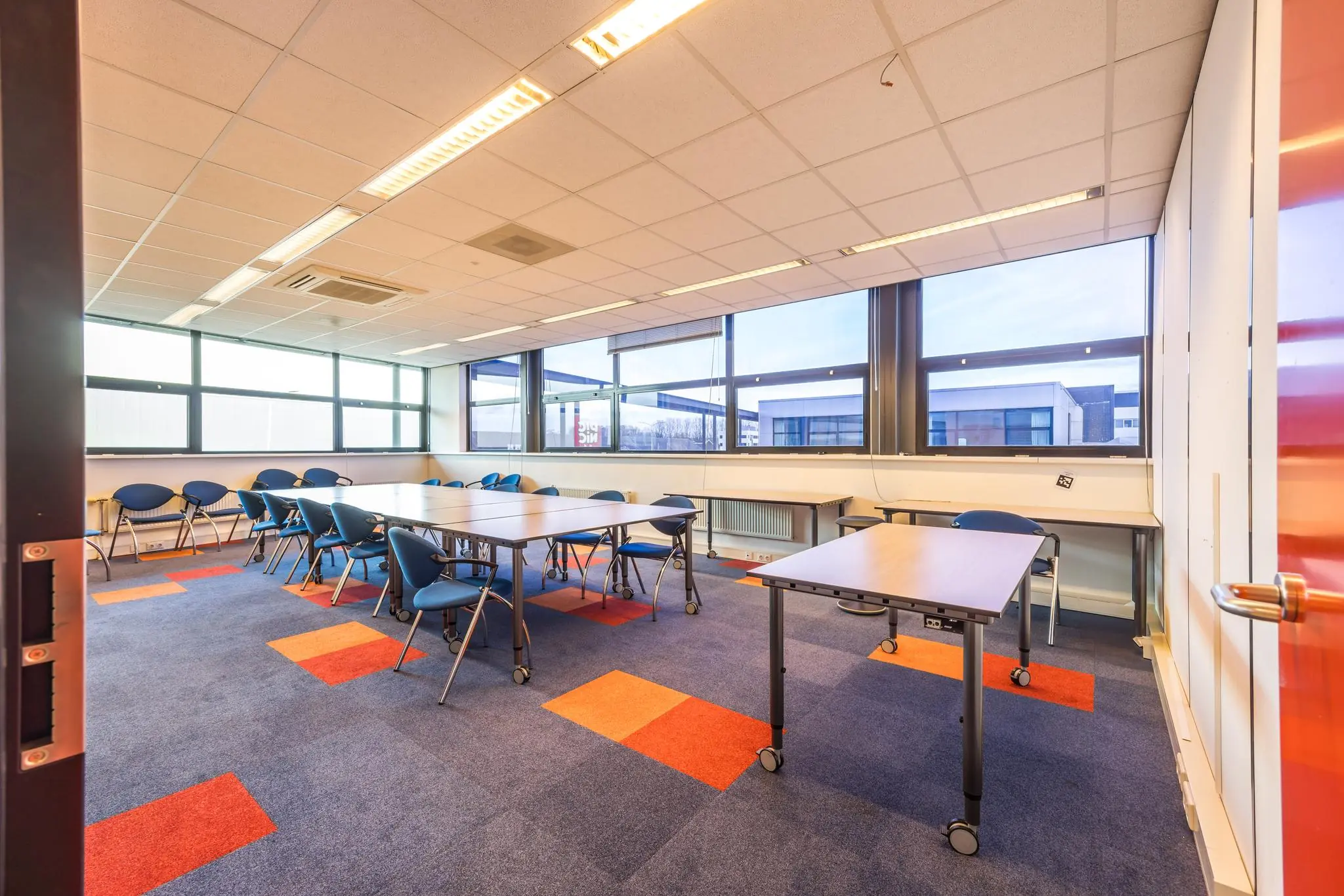 Vergaderruimte aan de Atoomweg met opklapbare tafels, blauwe stoelen en grote ramen met uitzicht op kantoorgebouwen.