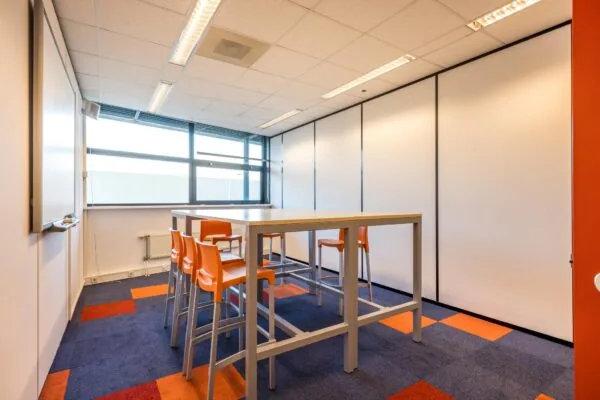 Vergaderruimte met hoge tafel, oranje stoelen en kleurrijk tapijt in een modern kantoor aan de Atoomweg.