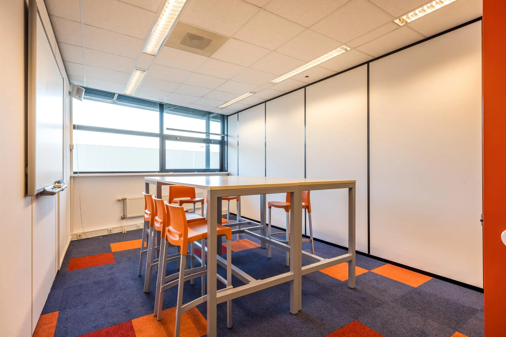 Vergaderruimte met hoge tafel, oranje stoelen en kleurrijk tapijt in een modern kantoor aan de Atoomweg.