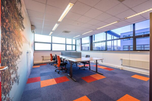 Moderne kantoorruimte aan de Atoomweg met kleurrijk tapijt, bureaus, oranje bureaustoelen en grote ramen voor natuurlijk licht.