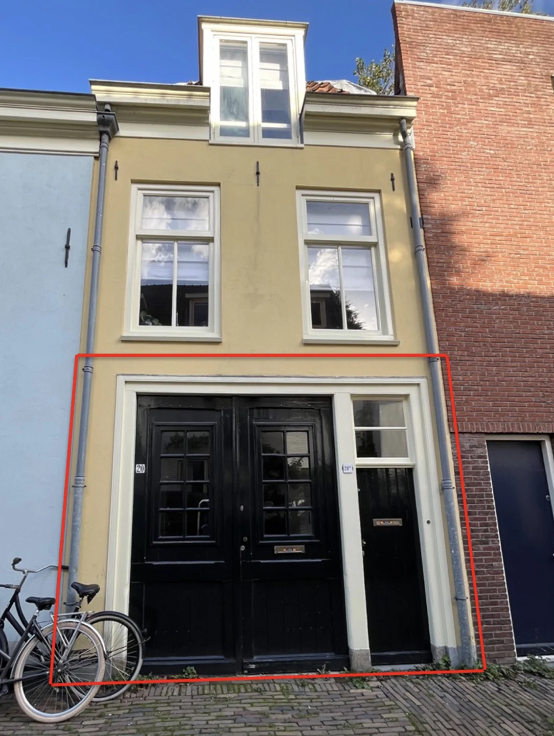 Voorgevel van een karakteristiek, geel geschilderd huis aan de Nobeldwarsstraat met zwarte deuren en fietsen ervoor.