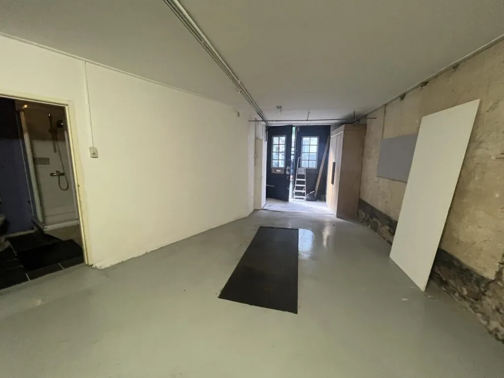 Een smalle, lege ruimte met betonvloer en zicht op een douchecabine links en een open deur met ladder aan het einde.