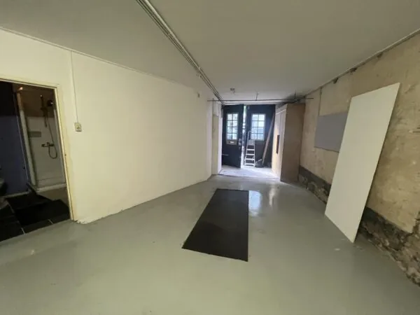 Een smalle, lege ruimte met betonvloer en zicht op een douchecabine links en een open deur met ladder aan het einde.