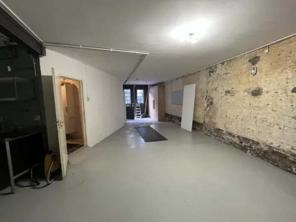 Een lege, langwerpige ruimte aan de Nobeldwarsstraat met een afgebladderde muur, een deur naar een andere kamer en zicht op een dubbele achterdeur met ladder.