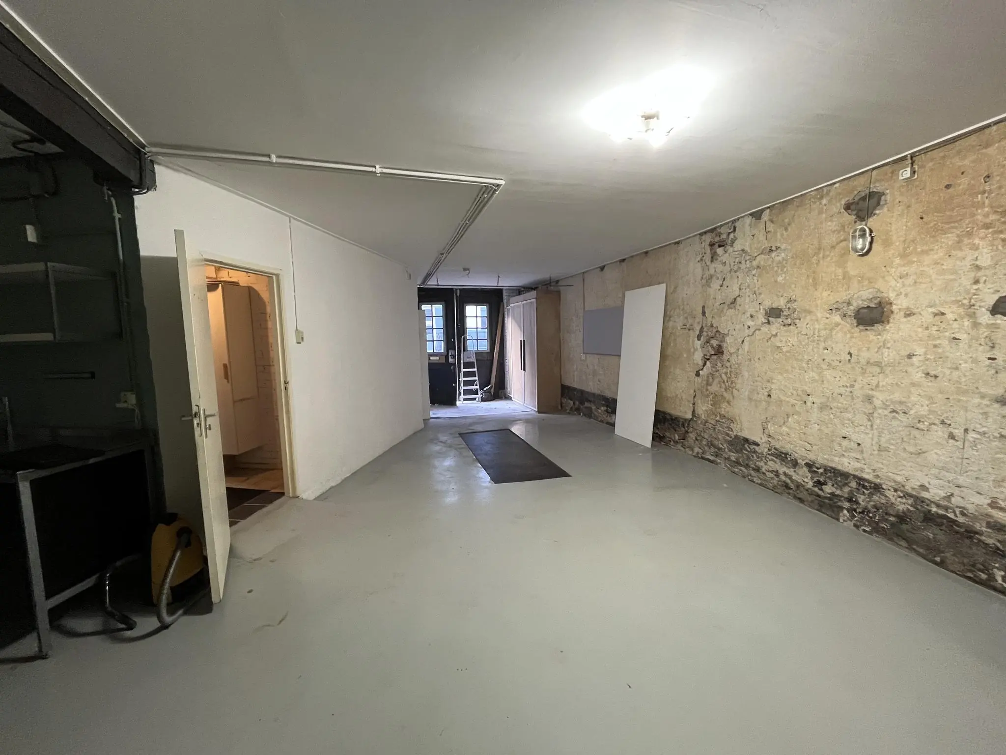 Een lege, langwerpige ruimte aan de Nobeldwarsstraat met een afgebladderde muur, een deur naar een andere kamer en zicht op een dubbele achterdeur met ladder.