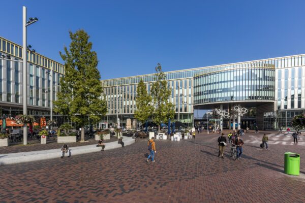 Het Hoekenrodeplein in Amsterdam-Zuidoost met moderne kantoorgebouwen, terrassen en voorbijgangers op een zonnige dag.