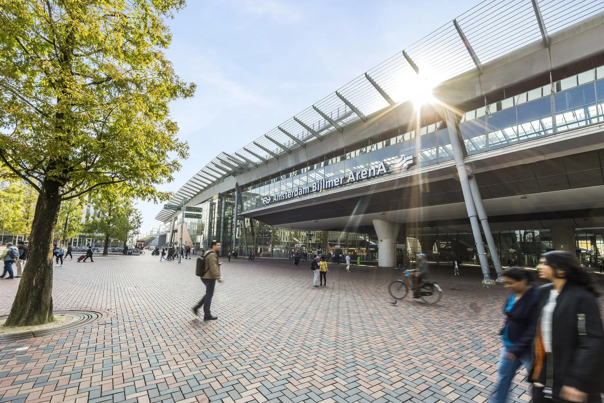 Vooraanzicht van station Amsterdam Bijlmer ArenA met lopende en fietsende mensen op het plein.