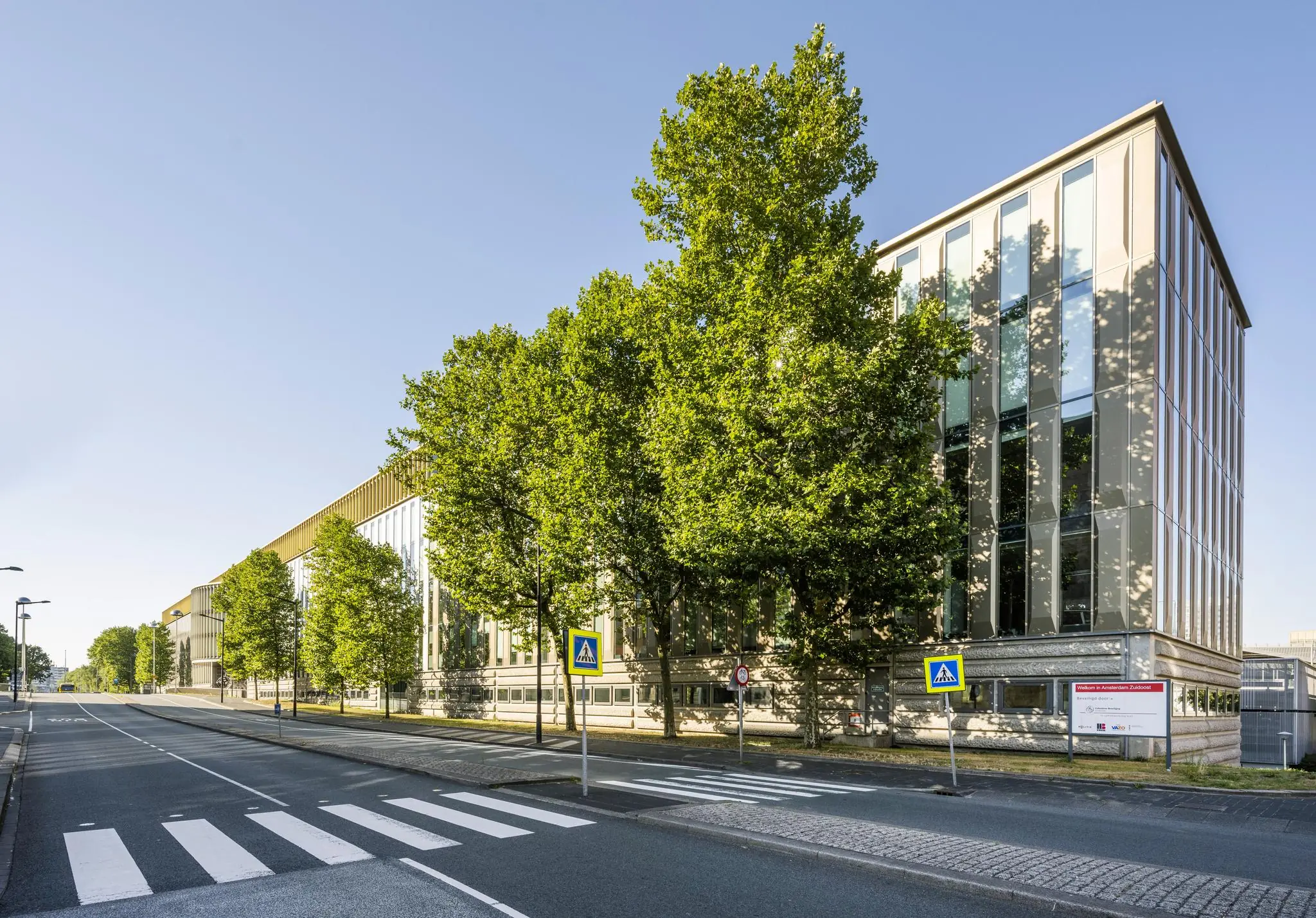 Modern kantoorgebouw aan de Hoekenrode in Amsterdam met bomen en een zebrapad op de voorgrond.